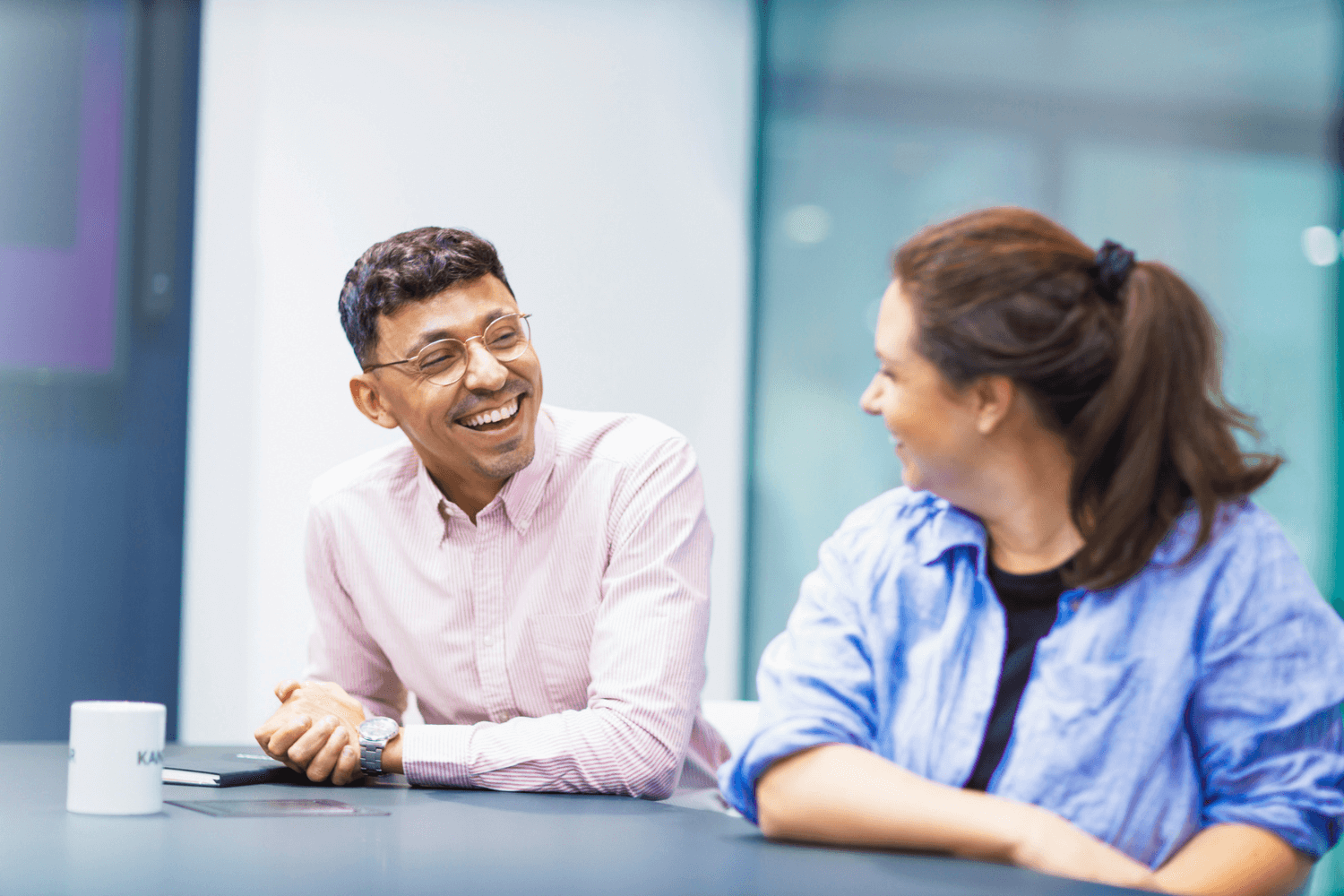 A man in a light pink striped shirt and glasses, sat at a table. He is smiling at the woman sat next to him. She is wearing a light blue short and has dark brown hair in a pony tail.