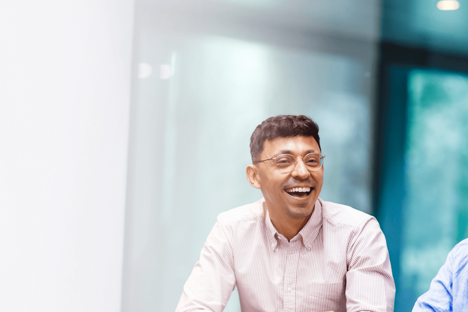 A young man with glasses with a light pink shirt, laughing and smiling at something off screen.