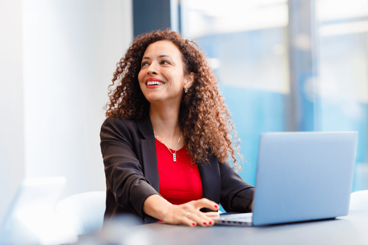 A woman in a black jacket and red top. She has long curly brown hair and is working in front of her laptop. She has been distracted by someone behind the camera.