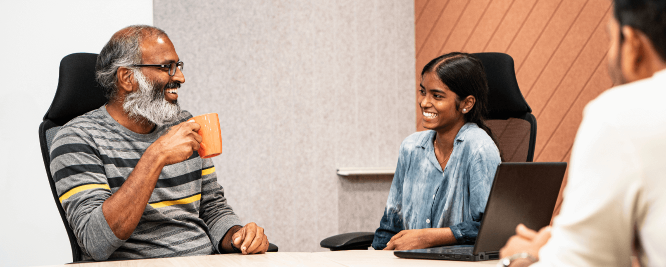 Dois colegas estão conversando juntos. À esquerda está um senhor mais velho. Ele é indiano, tem uma barba grisalha e está vestindo um suéter cinza com listras pretas e amarelas. Ele está bebendo de uma caneca laranja. Ele está conversando com uma colega mais jovem. Ela está vestindo uma blusa azul e tem o cabelo preso.