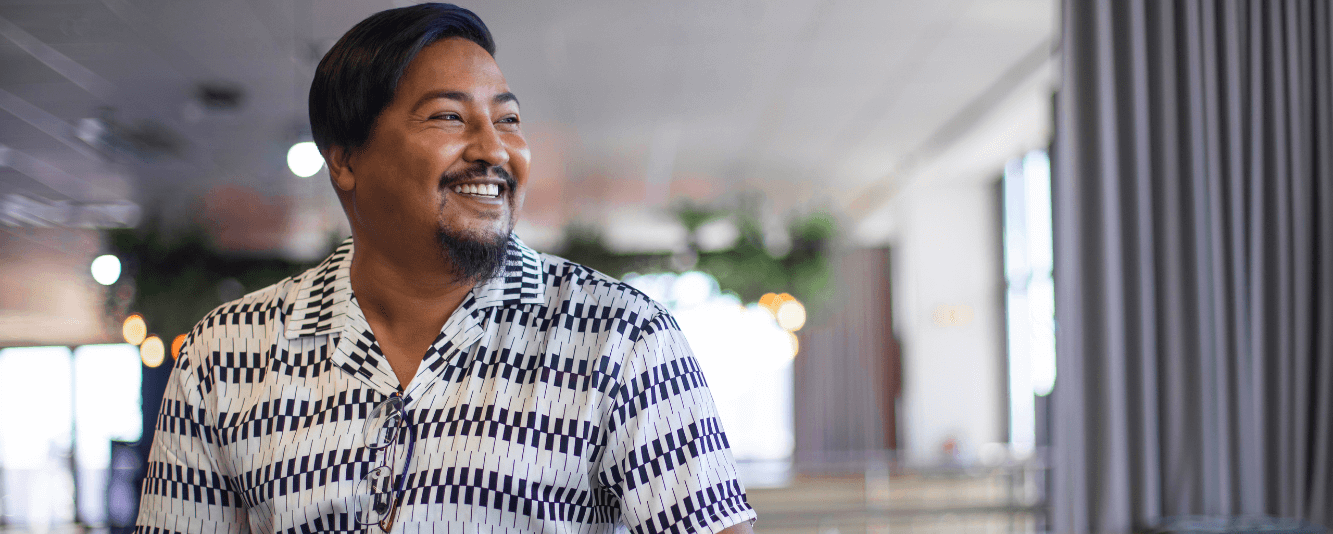 Um homem está sorrindo para a direita. Ele tem cabelo curto e castanho e está vestindo uma camisa preta e branca com um padrão que se assemelha a teclas de piano.