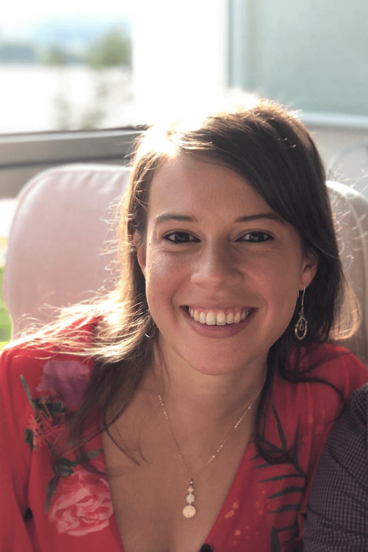 Uma mulher com cabelos escuros um pouco acima do comprimento dos ombros, sorrindo para a câmera e vestindo uma blusa vermelha com estampa floral. 