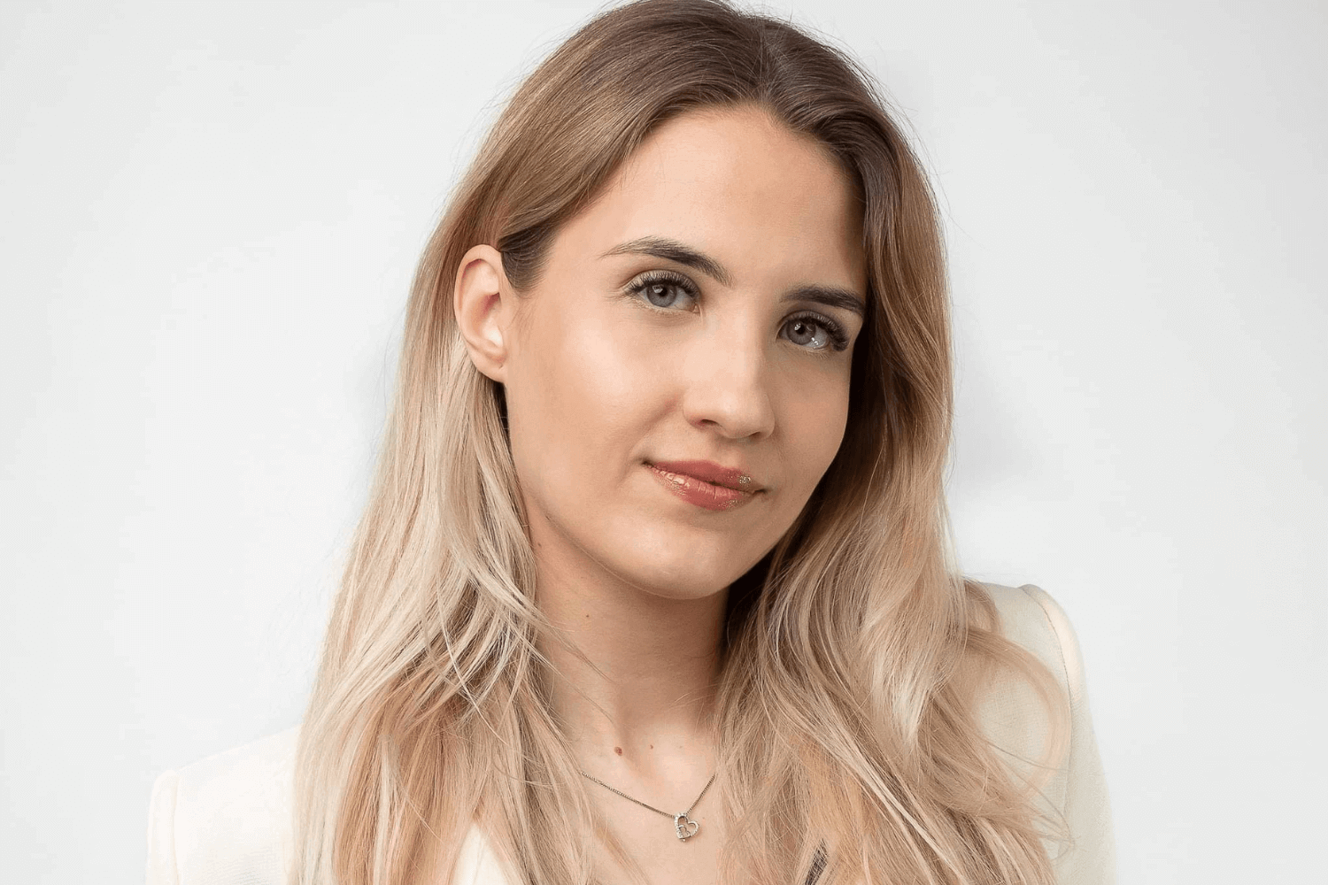 Uma mulher com longos cabelos loiros, vestindo um casaco branco com botões e uma camisola vermelha por baixo. 