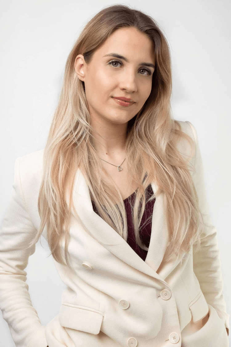 Uma mulher com longos cabelos loiros, vestindo um casaco branco com botões e uma camisola vermelha por baixo. 