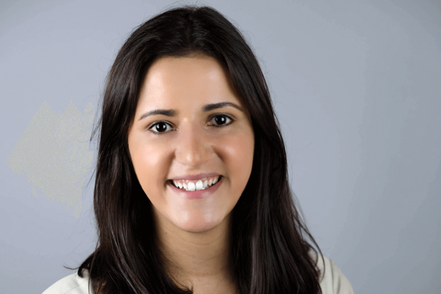 Uma mulher sorrindo para a câmera com os braços cruzados. Ela está vestindo uma jaqueta creme e tem cabelos castanhos na altura dos ombros.