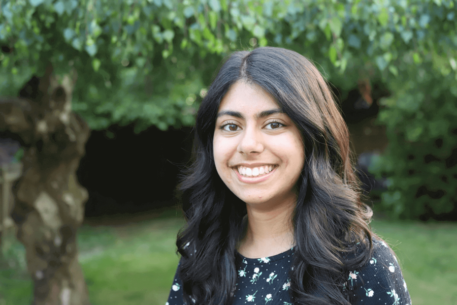 Uma mulher jovem com longos cabelos castanhos. Ela está vestindo uma blusa azul de mangas curtas com detalhes florais.
