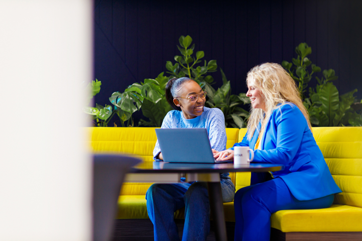 Zwei Frauen arbeiten gemeinsam vor einem Laptop an einem Tisch, der auf einem großen gelben Sofa steht. Eine Frau trägt einen hellblauen Pullover und Jeans. Die andere trägt einen blauen Anzug.