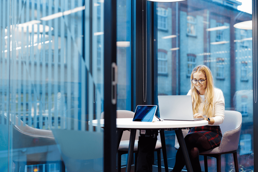 Eine blonde Frau arbeitet allein an ihrem Laptop in einem Besprechungsraum. Sie trägt ein weißes Oberteil und eine Brille.