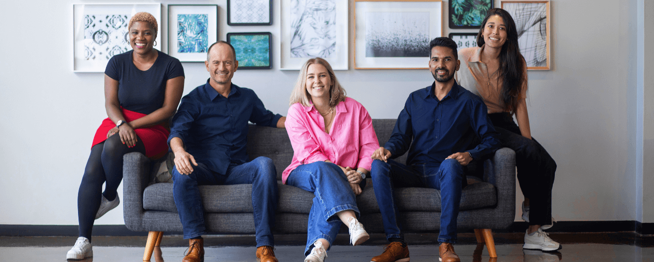 Eine Gruppe von fünf Personen sitzt auf einem grauen Sofa, mit einer Reihe von abstrakten Bildern in Rahmen hinter ihnen. Von links nach rechts. Eine schwarze Frau sitzt auf der Armlehne des Sofas. Sie trägt ein schwarzes T-Shirt und einen roten Rock. Als nächstes ein weißer Mann, der blaue Jeans und ein marineblaues Hemd trägt. In der Mitte eine blonde Frau, die ein leuchtend rosa Hemd, Jeans und weiße Turnschuhe trägt. Viertens ein weiterer Mann, der ebenfalls ein marineblaues Hemd und blaue Jeans trägt. Auf der anderen Armlehne des Sofas sitzt eine Frau in schwarzen Jeans, weißen Turnschuhen und einem braunen Oberteil.