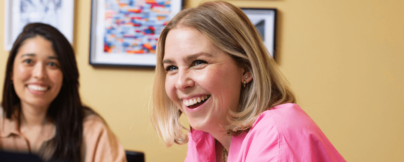 Eine Frau mit kurzen blonden Haaren und einem rosa Hemd lacht in die Kamera. Sie sitzt in einem Raum, der hellgelb gestrichen ist. Hinter ihr ist eine andere weibliche Kollegin, leicht unscharf. Sie hat lange braune Haare und trägt ein braunes Oberteil.