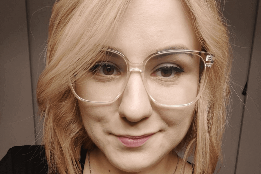 Eine Frau mit einem schwarzen Oberteil und einer Brille mit durchsichtigem Gestell. Sie hat schulterlanges, blondes Haar.