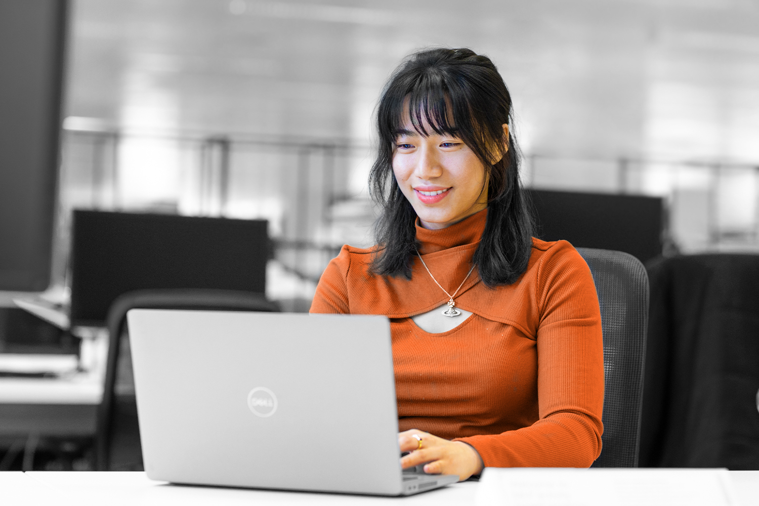 Imagen en blanco y negro, en la que sólo la ropa y el tono de la piel aparecen en color. Una mujer joven con un top naranja trabaja con un ordenador portátil. 