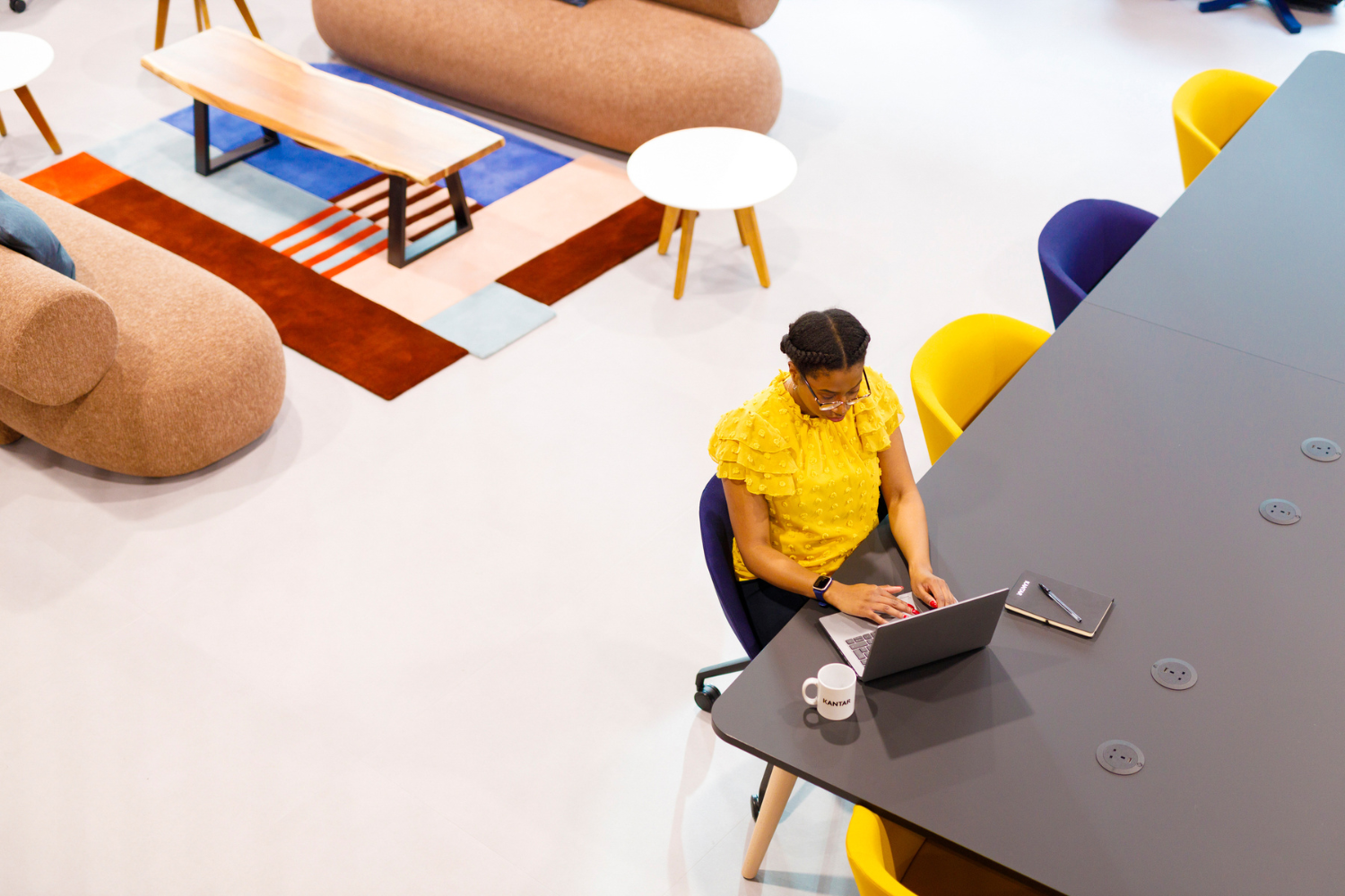 Una mujer trabaja con un portátil en una mesa larga. Lleva un top amarillo. La foto está tomada desde arriba. 