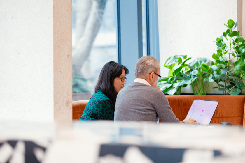 Dos colegas trabajan juntos desde un ordenador portátil. Ella tiene el pelo oscuro hasta los hombros, lleva gafas y un vestido verde. Él lleva un jersey gris y una camisa blanca.