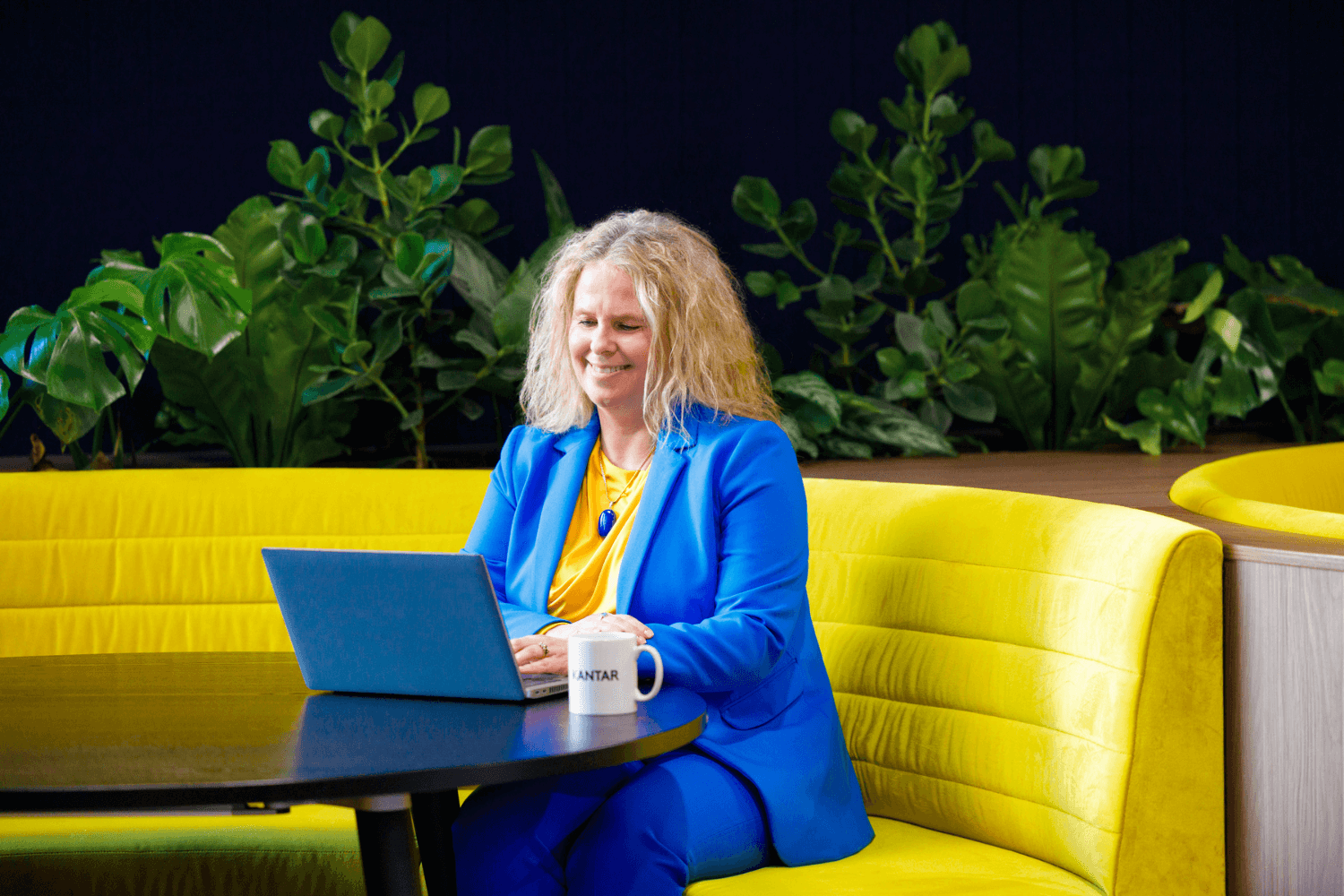 Una mujer de larga melena rubia rizada trabajaba con un portátil en un sofá amarillo. 