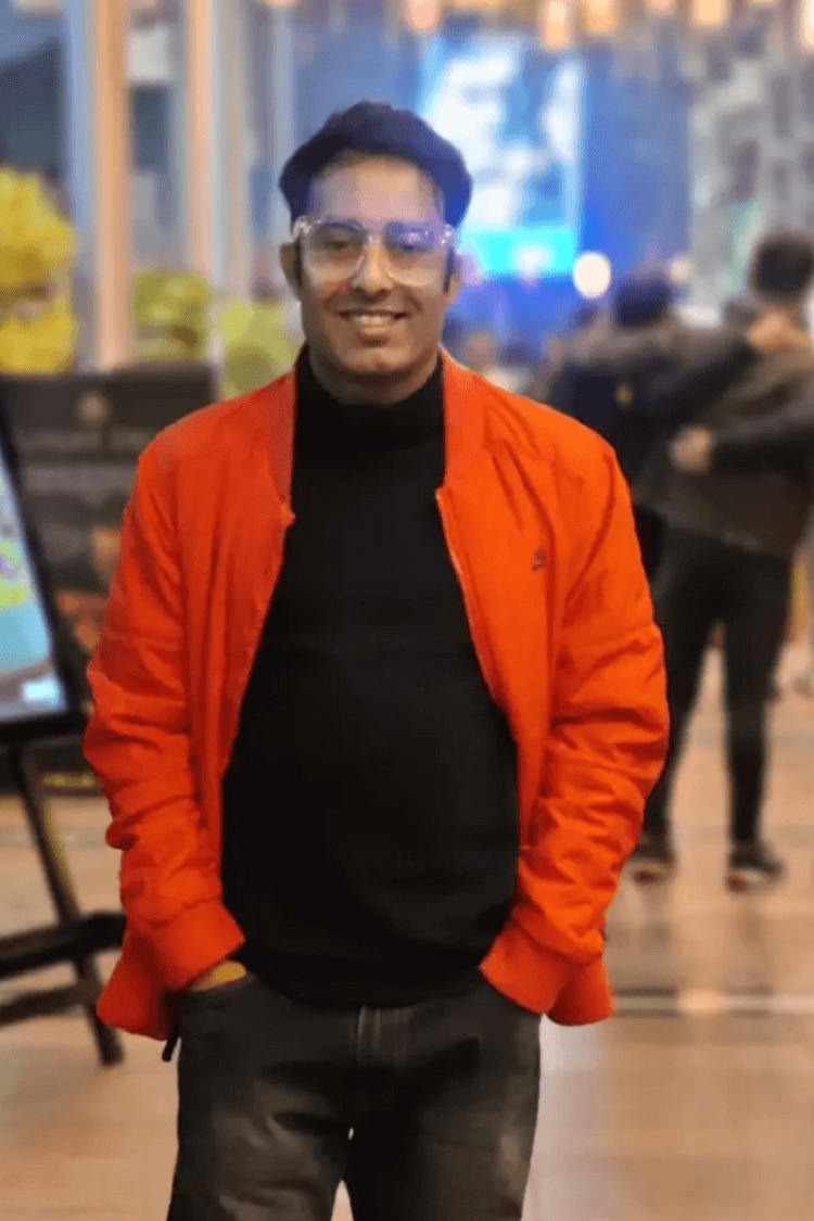 Un hombre con una chaqueta naranja y un polo negro, sonriendo directamente a la cámara. Está en el exterior de una escena tipo cafetería. 