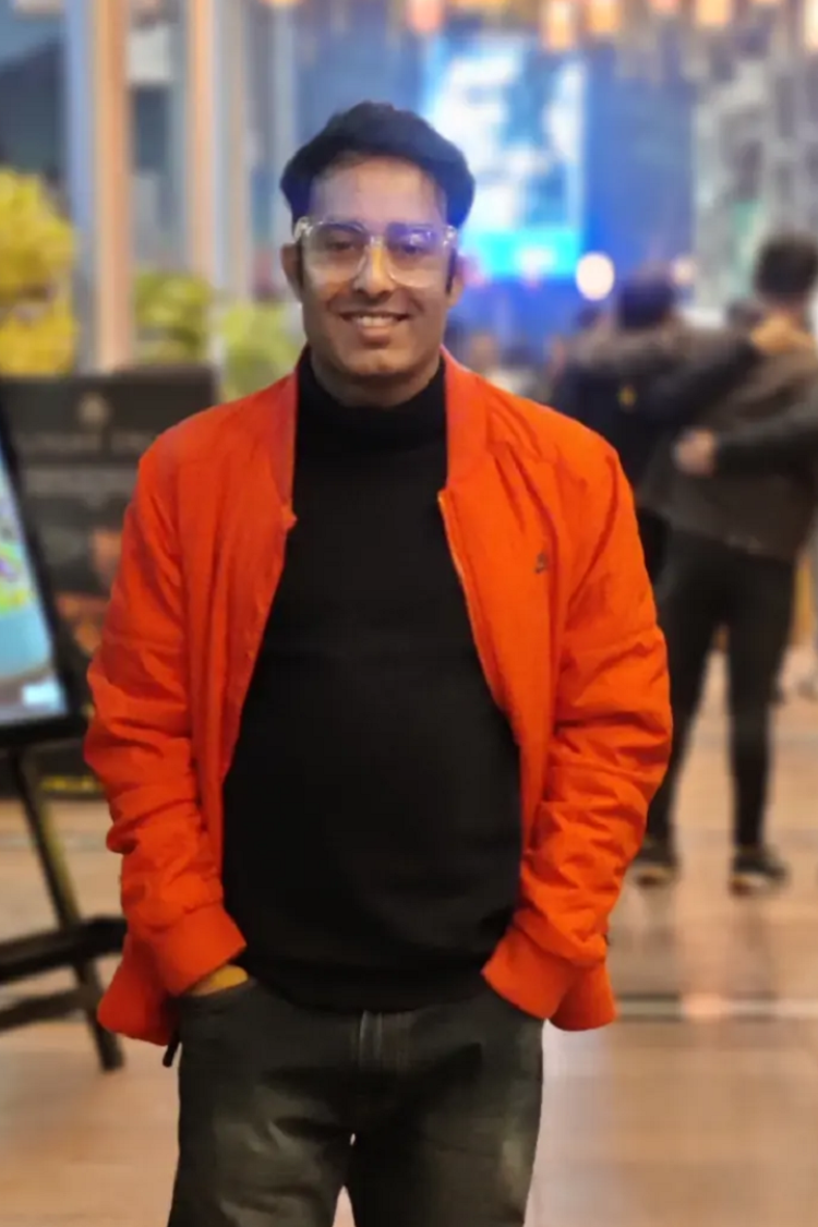 Un hombre con una chaqueta naranja y un polo negro, sonriendo directamente a la cámara. Está en el exterior de una escena tipo cafetería. 