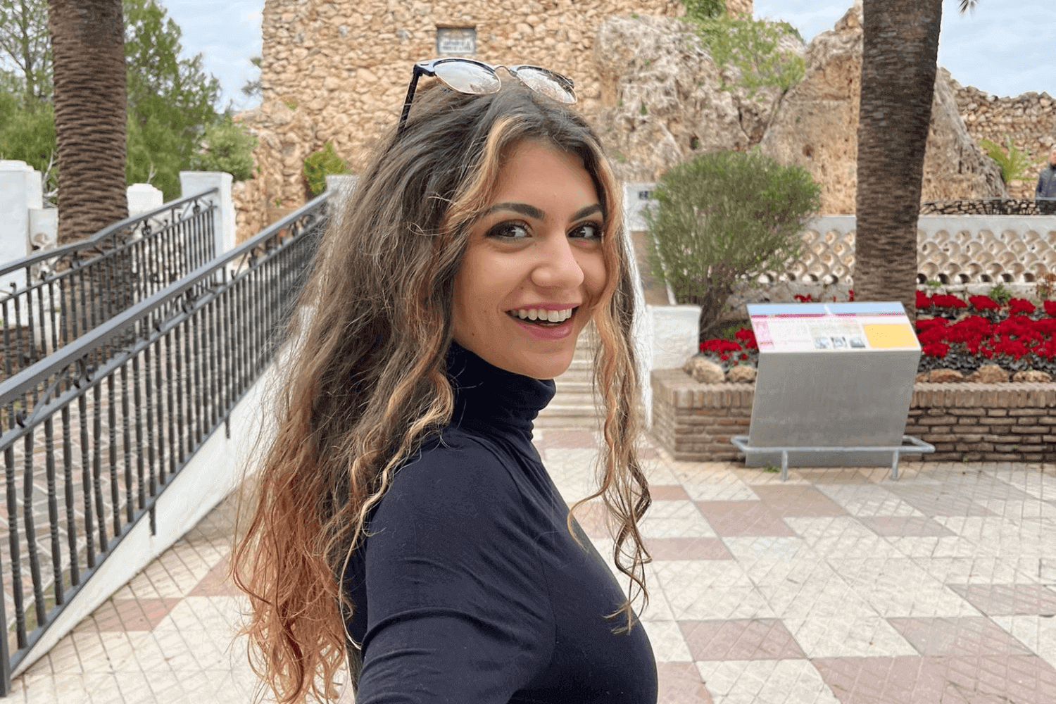 Una mujer joven mira a la cámara. Lleva el pelo rizado y gafas de sol en la cabeza. Lleva un polo negro. 