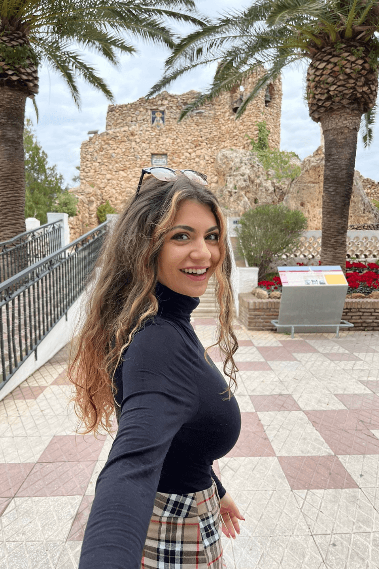 Una mujer joven mira a la cámara. Lleva el pelo rizado y gafas de sol en la cabeza. Lleva un polo negro. 