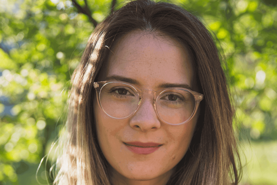 Una mujer con un polo negro, pelo largo castaño claro y gafas. Está al aire libre.