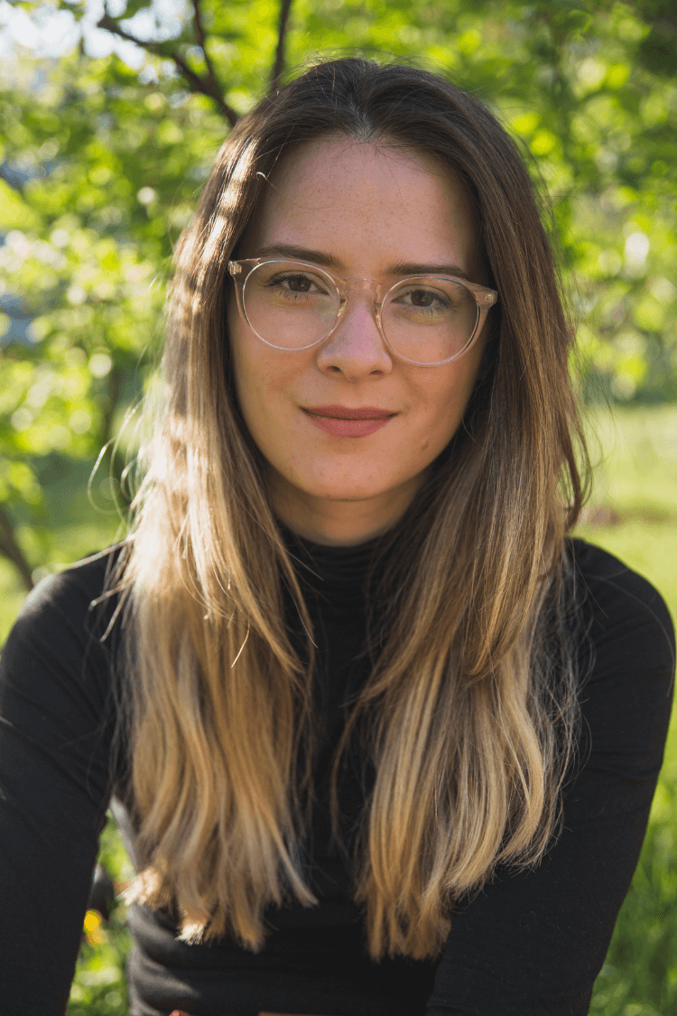 Una mujer con un polo negro, pelo largo castaño claro y gafas. Está al aire libre.