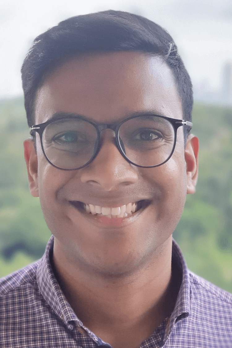 Un joven con gafas redondas de montura negra sonríe a la cámara. Lleva una camisa de cuadros y tiene el pelo corto y castaño.