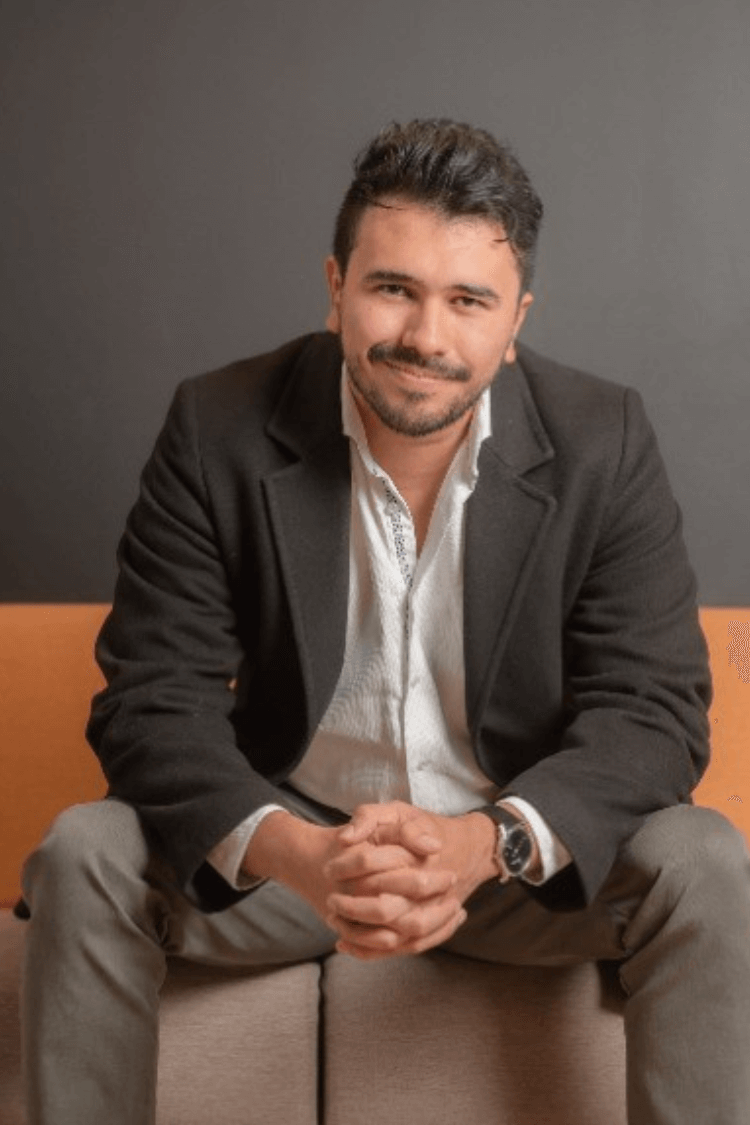 Un hombre sentado en un sofá naranja y marrón. Lleva pantalones chinos grises, camisa blanca y chaqueta gris.  