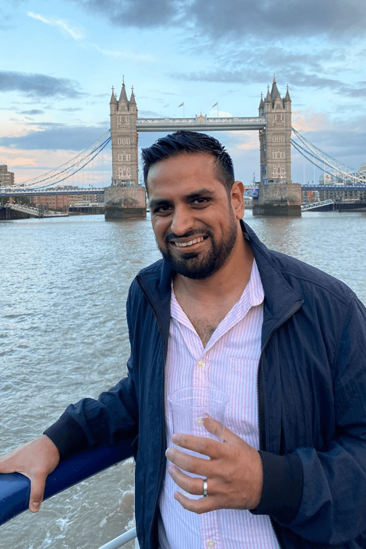 Un hombre con chaqueta azul y camisa clara. El Tower Bridge está detrás de él.