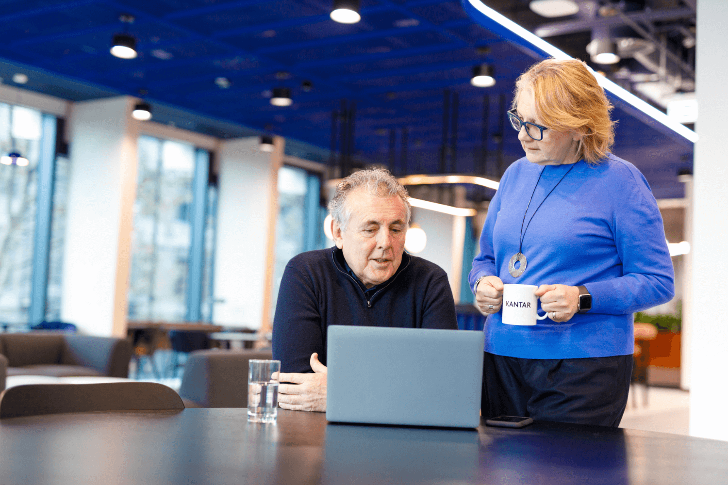 Une femme en pull bleu est debout et tient un café. A la table, un homme en pull noir, les bras croisés, regarde son ordinateur portable.