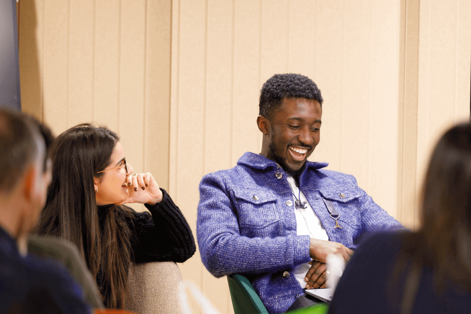 Un jeune homme en veste bleue et chemise blanche rit de quelque chose devant lui. Sa collègue à côté de lui le regarde avec la main sur la bouche.  