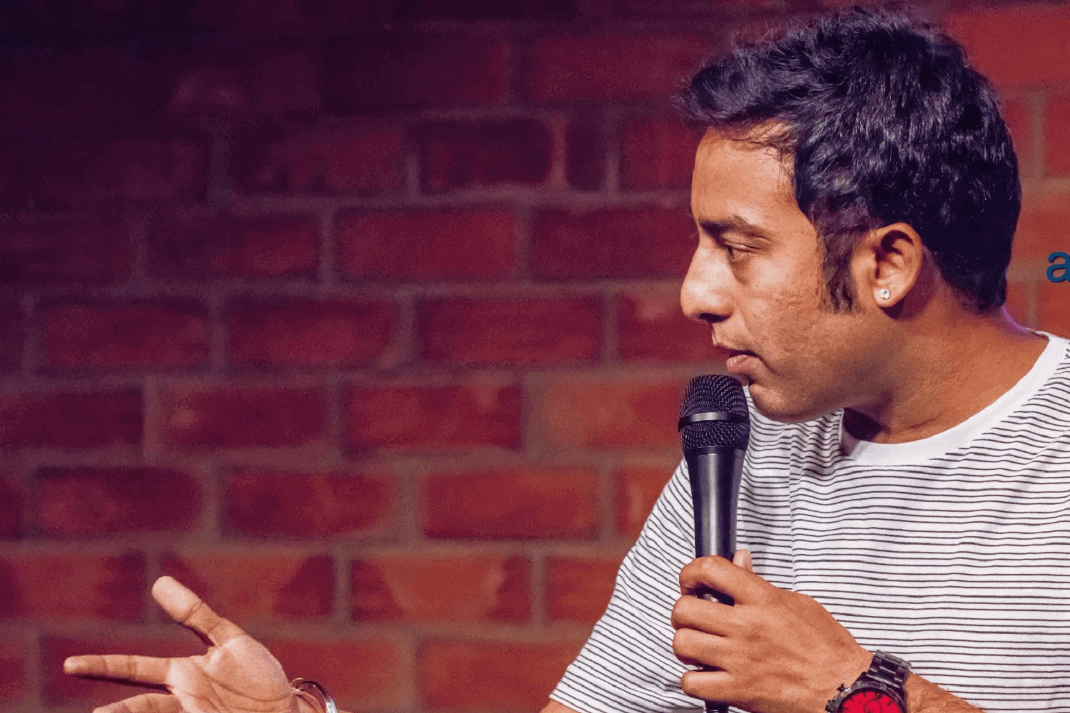 Un homme sur scène. Il porte un T-shirt à rayures horizontales gris et clair et tient un micro. 