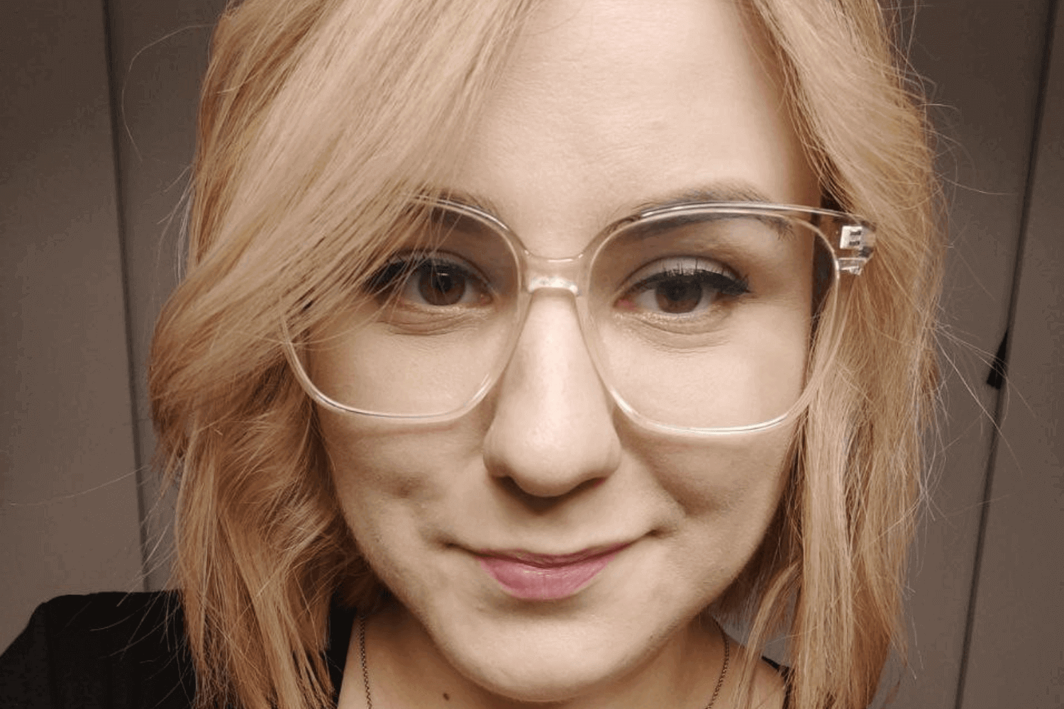 Une femme avec un haut noir et des lunettes à monture transparente. Elle a des cheveux blonds au niveau des épaules. 