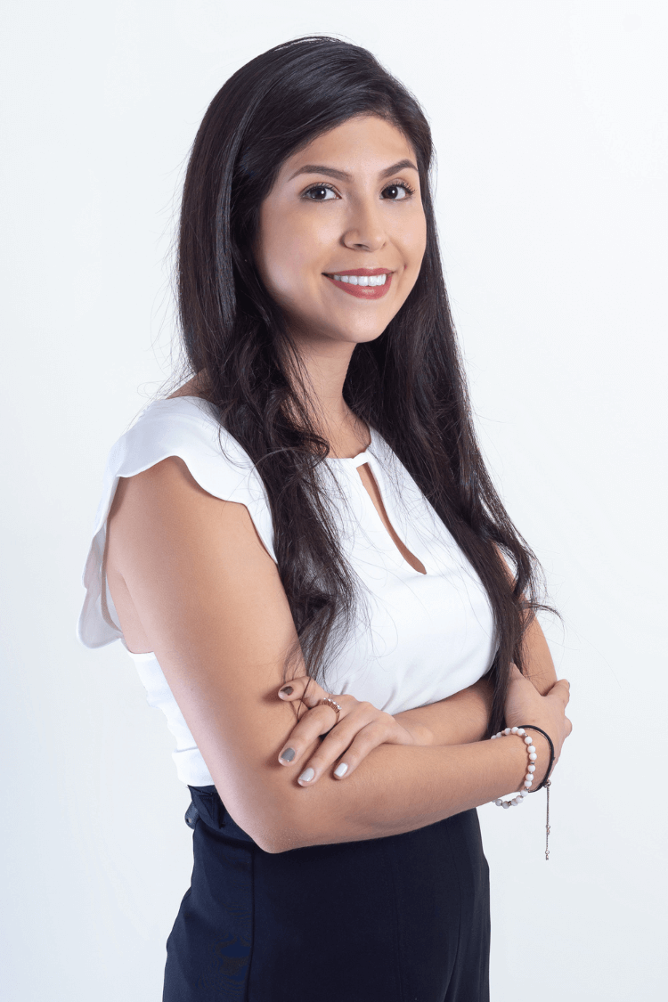 Une femme aux longs cheveux bruns. Elle a les bras croisés et porte une chemise blanche à manches courtes. 