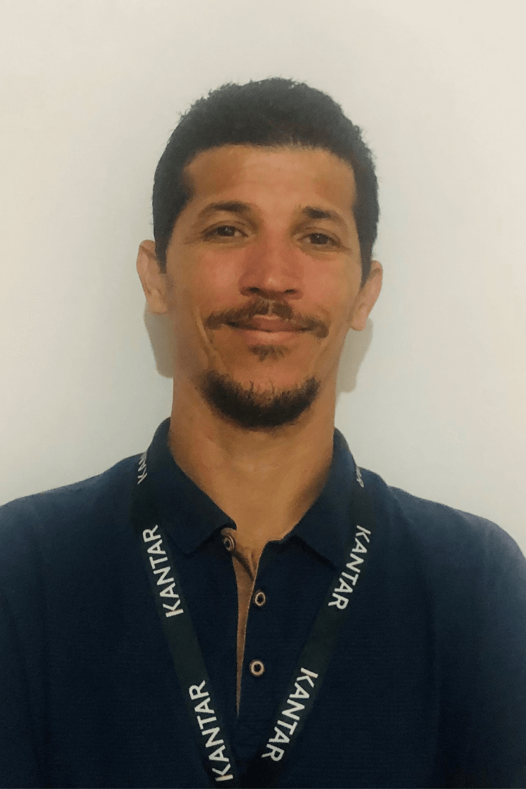 Un homme aux cheveux bruns et à la barbe brun foncé. Il porte une chemise bleu marine et on peut également voir son cordon Kantar.