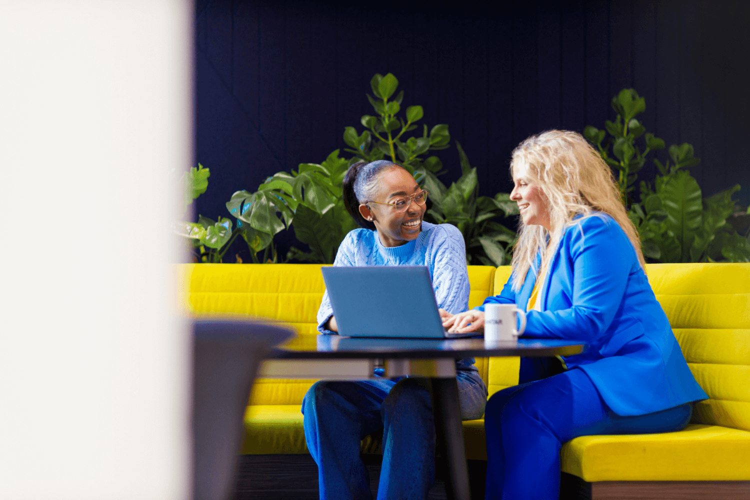 Twee vrouwen werken samen voor een laptop aan een tafel op een grote gele bank. De ene vrouw draagt een lichtblauwe trui en spijkerbroek. De andere draagt een blauw pak.