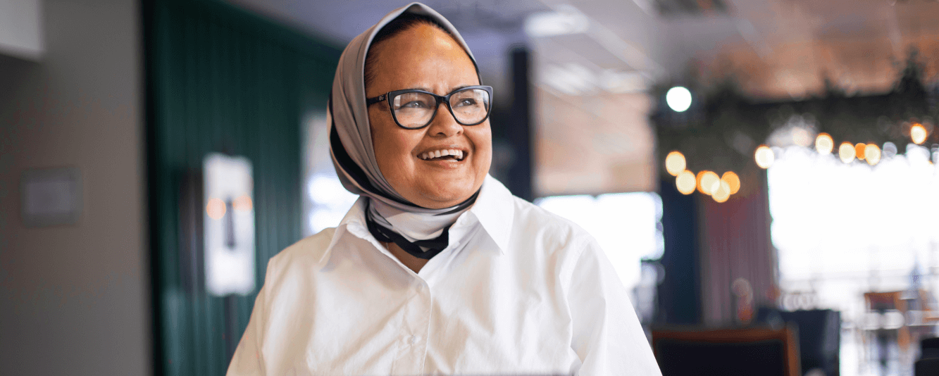 Een vrouw in een wit shirt glimlacht naar rechts. Ze draagt een hoofddoek, die een mix is van lichte en donkere kleuren. Ze draagt een bril.
