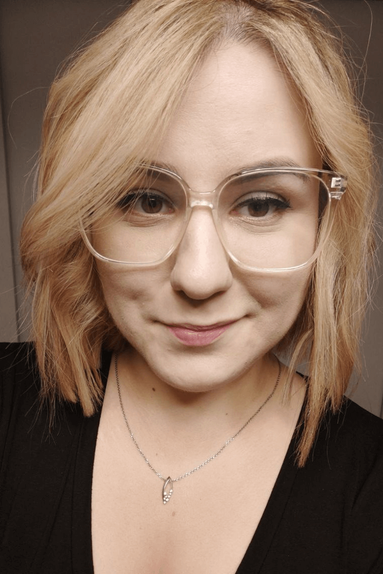 Een vrouw met een zwart topje en een bril met doorzichtig montuur. Ze heeft schouderlang blonder haar.