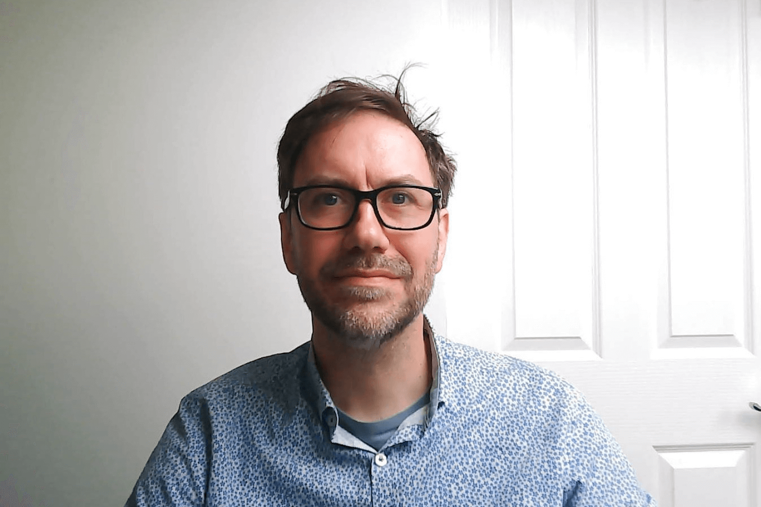 Een man met een bril met zwarte rand kijkt naar de camera, met een blauw shirt aan. Hij heeft kort bruin haar. 