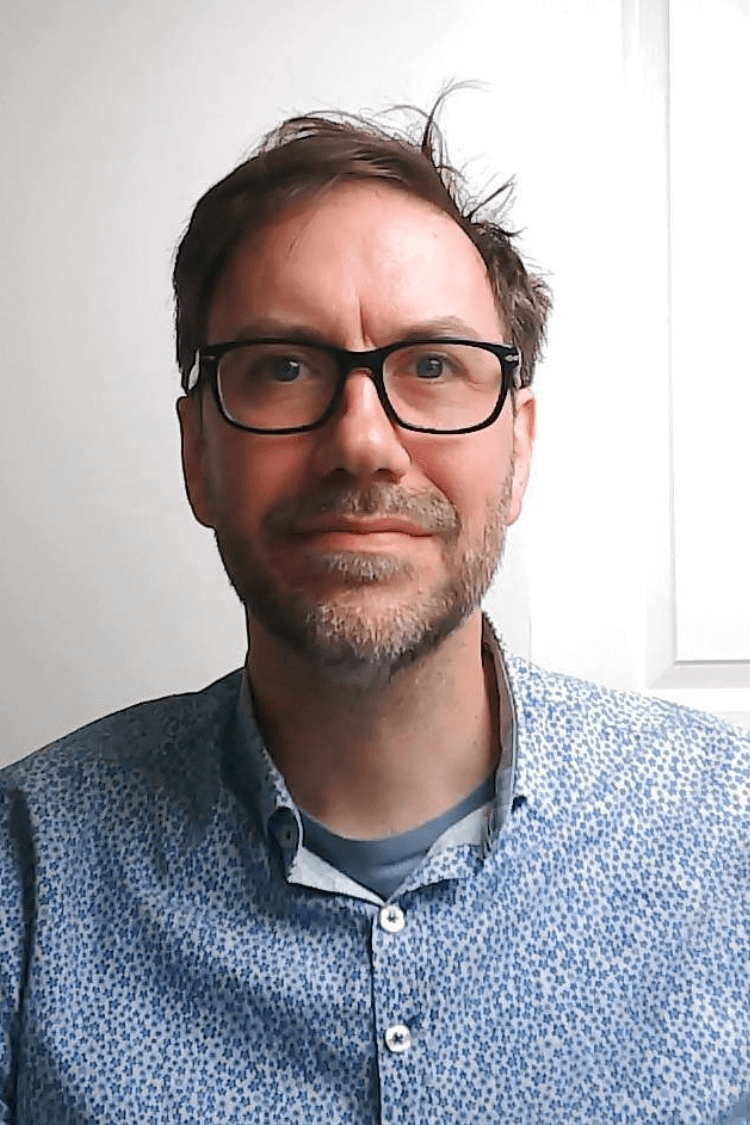 Een man met een bril met zwarte rand kijkt naar de camera, met een blauw shirt aan. Hij heeft kort bruin haar.