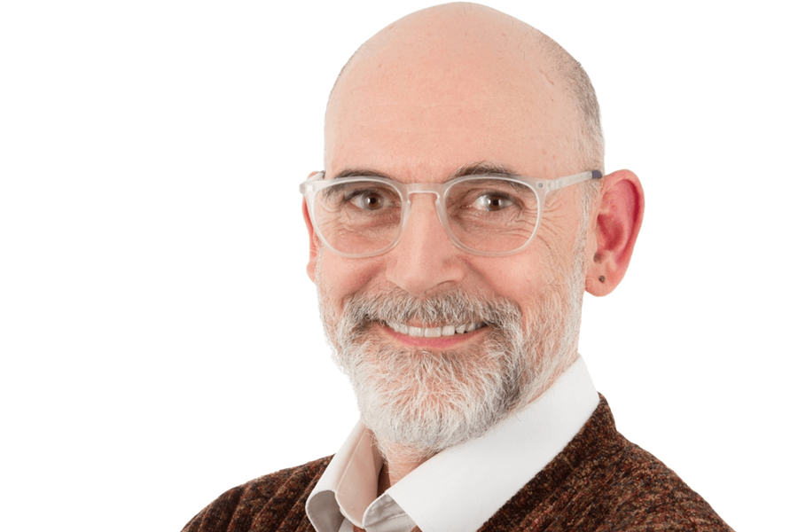 Een heer met een baard en een bril. Hij draagt een bruine trui, wit overhemd en stropdas.