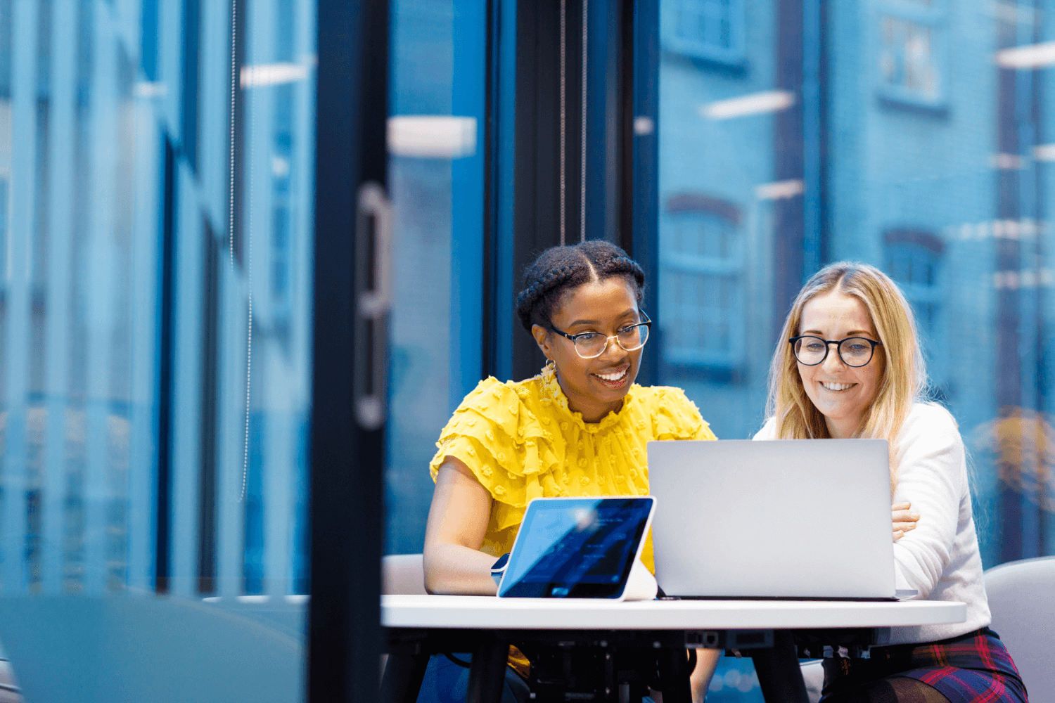 两位女士一起在会议室里使用笔记本电脑工作。其中一位穿着黄色上衣，另一位穿着白色毛衣。